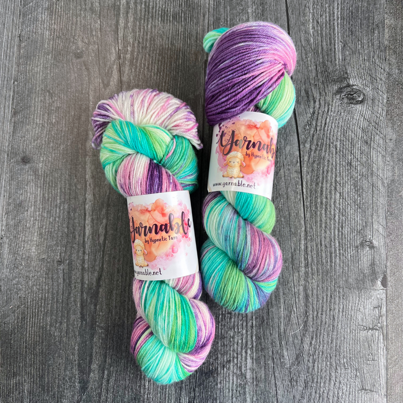 Hand-dyed yarn skeins featuring green, teal, purple, and pink color variations, displayed side by side on a gray wood background with Yarnable labels visible.