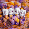 Four multicolored hand-dyed yarn skeins in warm orange and yellow with purple accents, photographed on a textured backdrop made of coordinating yarn.