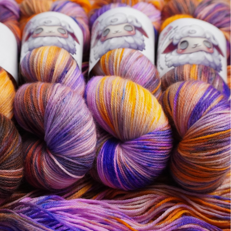 Four multicolored hand-dyed yarn skeins in warm orange and yellow with purple accents, photographed on a textured backdrop made of coordinating yarn.