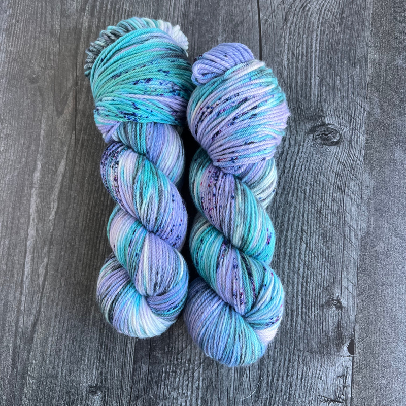 Several twisted yarn skeins in cool teal, turquoise, and lavender shades with speckled details, displayed on a textured yarn backdrop.