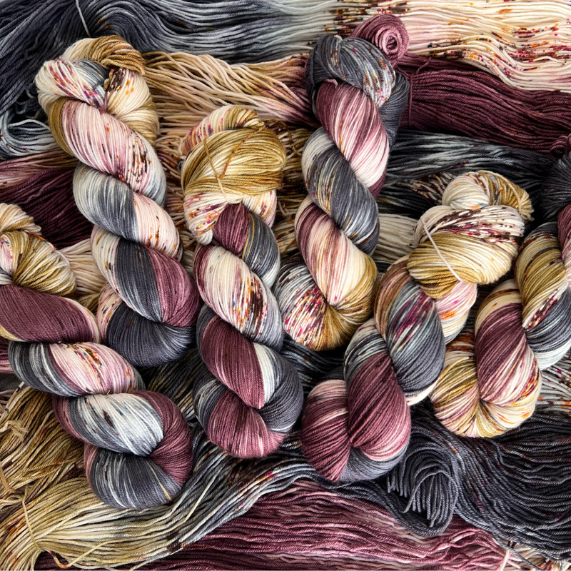 Close-up of multiple twisted skeins of hand-dyed yarn featuring burgundy, charcoal gray, ivory, and mustard color variations.