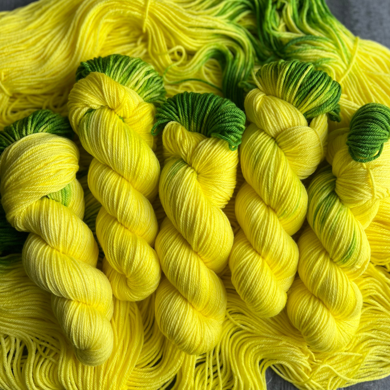 Hand-dyed yarn skeins in bright yellow with green accents, twisted and arranged on a matching yellow yarn background.