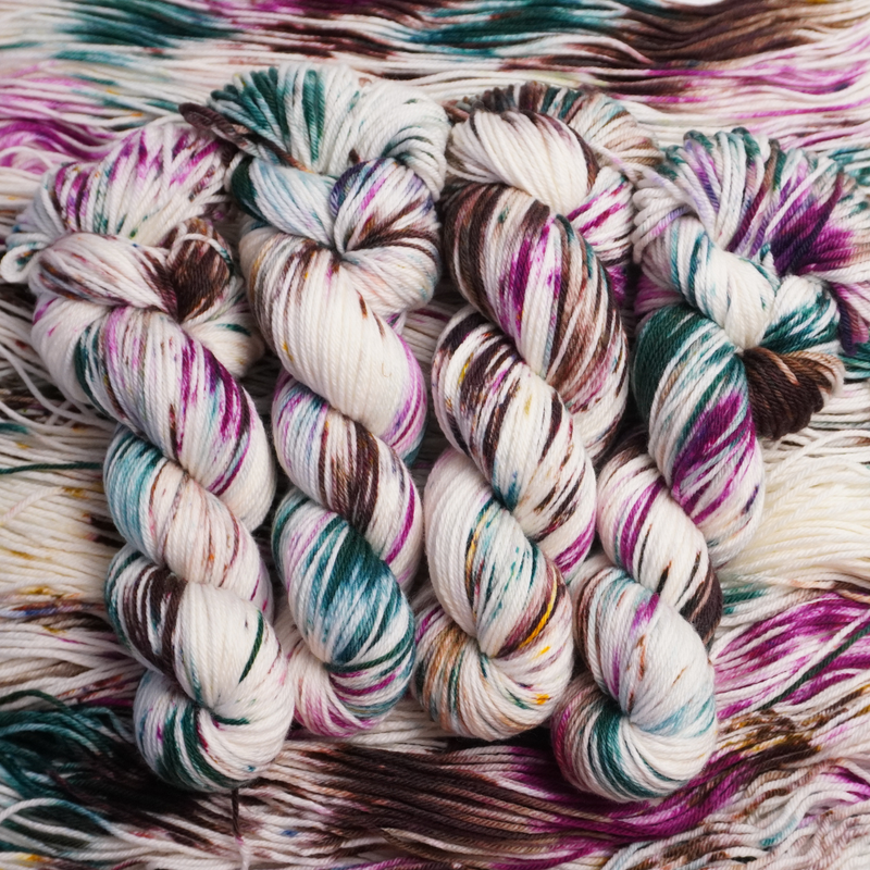 Twisted skeins of hand-dyed yarn with multicolored streaks of teal, pink, brown, and green on a light base, displayed on loose yarn.