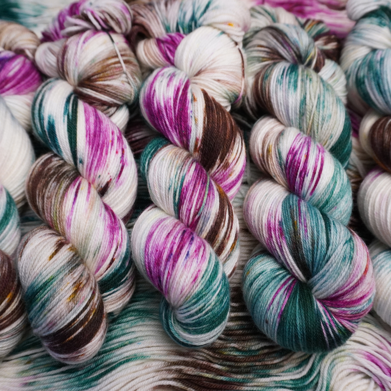 Twisted skeins of hand-dyed yarn with multicolored streaks of teal, pink, brown, and green on a light base, displayed on loose yarn.