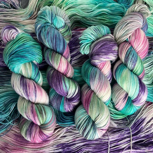 Hand-dyed yarn skeins featuring green, teal, purple, and pink color variations, displayed side by side on a gray wood background with Yarnable labels visible.