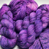 Bright purple speckled Zodiac Sagittarius yarn skeins and a yarn cake piled on purple knit swatches