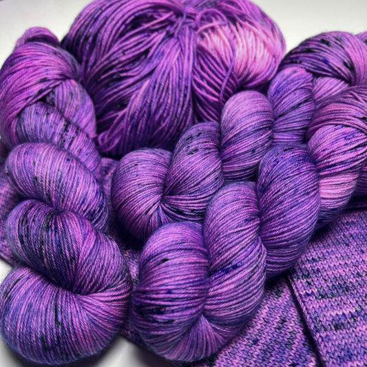 Bright purple speckled Zodiac Sagittarius yarn skeins and a yarn cake piled on purple knit swatches