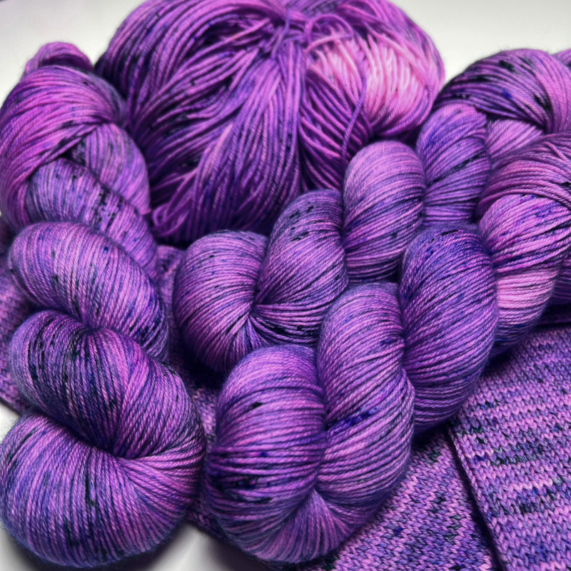 Bright purple speckled Zodiac Sagittarius yarn skeins and a yarn cake piled on purple knit swatches
