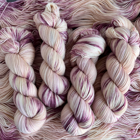 Hand-dyed yarn skeins in soft cream, blush pink, and mauve tones, twisted and arranged side by side with subtle variegation.