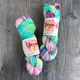 Hand-dyed yarn skeins featuring green, teal, purple, and pink color variations, displayed side by side on a gray wood background with Yarnable labels visible.