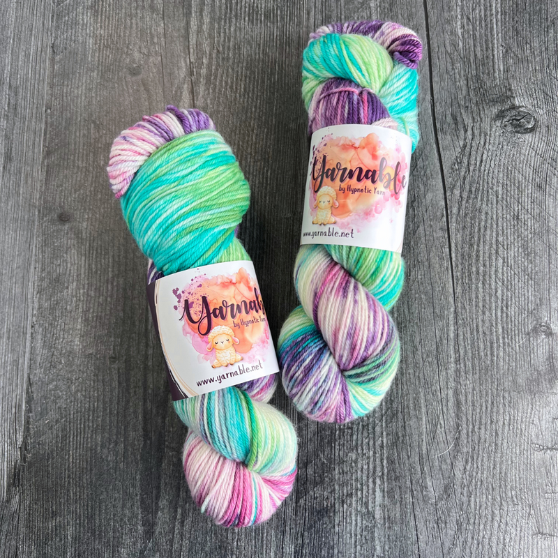Hand-dyed yarn skeins featuring green, teal, purple, and pink color variations, displayed side by side on a gray wood background with Yarnable labels visible.