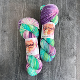 Hand-dyed yarn skeins featuring green, teal, purple, and pink color variations, displayed side by side on a gray wood background with Yarnable labels visible.