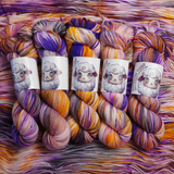Four multicolored hand-dyed yarn skeins in warm orange and yellow with purple accents, photographed on a textured backdrop made of coordinating yarn.