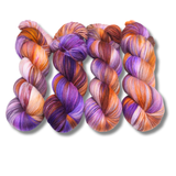 Four multicolored hand-dyed yarn skeins in warm orange and yellow with purple accents, photographed on a textured backdrop made of coordinating yarn.