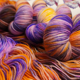 Four multicolored hand-dyed yarn skeins in warm orange and yellow with purple accents, photographed on a textured backdrop made of coordinating yarn.