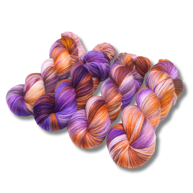Four multicolored hand-dyed yarn skeins in warm orange and yellow with purple accents, photographed on a textured backdrop made of coordinating yarn.