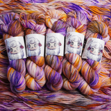 Four multicolored hand-dyed yarn skeins in warm orange and yellow with purple accents, photographed on a textured backdrop made of coordinating yarn.