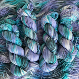 Several twisted yarn skeins in cool teal, turquoise, and lavender shades with speckled details, displayed on a textured yarn backdrop.
