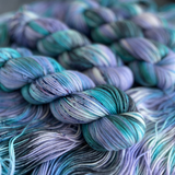 Several twisted yarn skeins in cool teal, turquoise, and lavender shades with speckled details, displayed on a textured yarn backdrop.