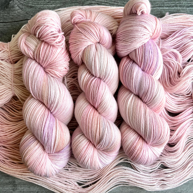 Hand-dyed yarn skeins in soft pastel pink with subtle lavender and cream variations, shown twisted and arranged side by side.