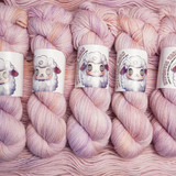 Hand-dyed yarn skeins in soft pastel pink with subtle lavender and cream variations, shown twisted and arranged side by side.