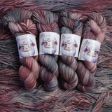Twisted skeins of hand-dyed yarn featuring blended plum, blush, and slate gray colors, shown with illustrated sheep labels.