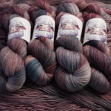 Twisted skeins of hand-dyed yarn featuring blended plum, blush, and slate gray colors, shown with illustrated sheep labels.