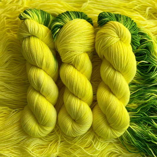 Hand-dyed yarn skeins in bright yellow with green accents, twisted and arranged on a matching yellow yarn background.