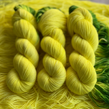 Hand-dyed yarn skeins in bright yellow with green accents, twisted and arranged on a matching yellow yarn background.