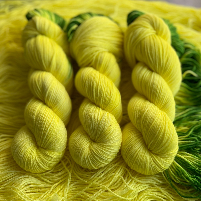 Hand-dyed yarn skeins in bright yellow with green accents, twisted and arranged on a matching yellow yarn background.