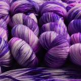 Hand-dyed yarn skeins in shades of purple, lavender, and soft pink, twisted and arranged on a matching purple yarn background.