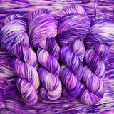 Hand-dyed yarn skeins in shades of purple, lavender, and soft pink, twisted and arranged on a matching purple yarn background.
