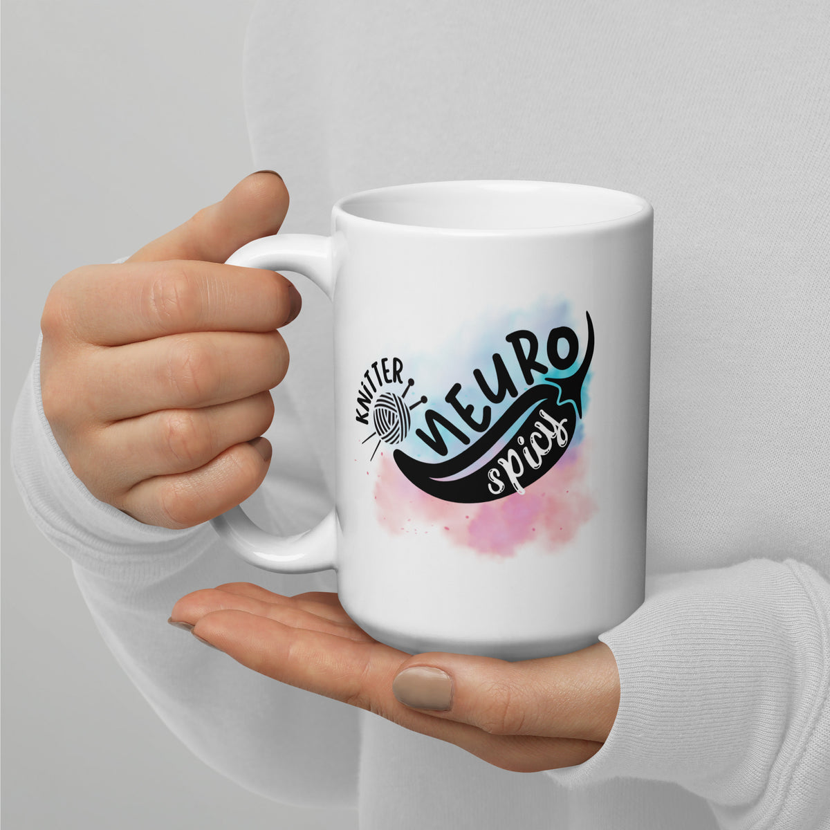 15 oz white mug with the NeuroSpicy Knitter design and a pink and blue watercolor splash, held in a hand