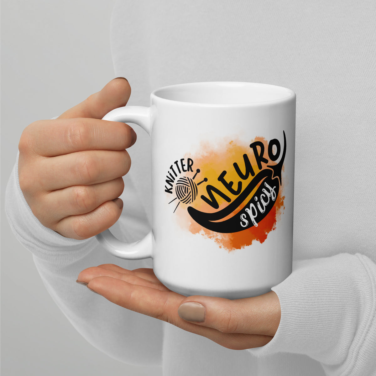 15 oz white mug with the NeuroSpicy Knitter design and an orange watercolor splash, held in a hand