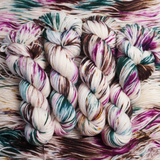 Twisted skeins of hand-dyed yarn with multicolored streaks of teal, pink, brown, and green on a light base, displayed on loose yarn.