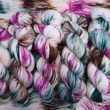 Twisted skeins of hand-dyed yarn with multicolored streaks of teal, pink, brown, and green on a light base, displayed on loose yarn.