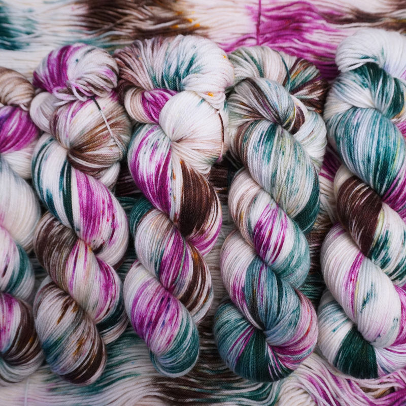 Twisted skeins of hand-dyed yarn with multicolored streaks of teal, pink, brown, and green on a light base, displayed on loose yarn.