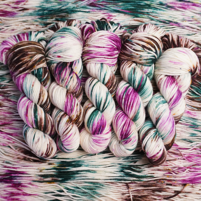 Twisted skeins of hand-dyed yarn with multicolored streaks of teal, pink, brown, and green on a light base, displayed on loose yarn.